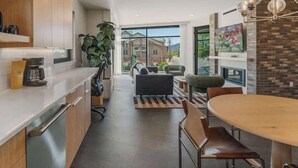 A modern kitchen with wooden cabinets, a dining area with a round table and chairs, and a large window offering a view of buildings outside.