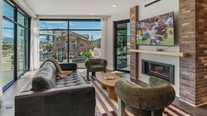 A modern living room with a leather sofa, armchairs, a fireplace, and a large TV mounted on a brick wall.