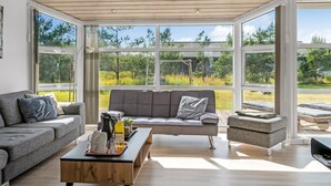 Una sala de estar moderna con un sofá gris, una mesa de centro de madera y vistas a un jardín a través de amplios ventanales.