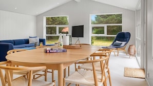 A modern living room with a wooden dining table, blue sofa, and a large window offering a view of greenery.
