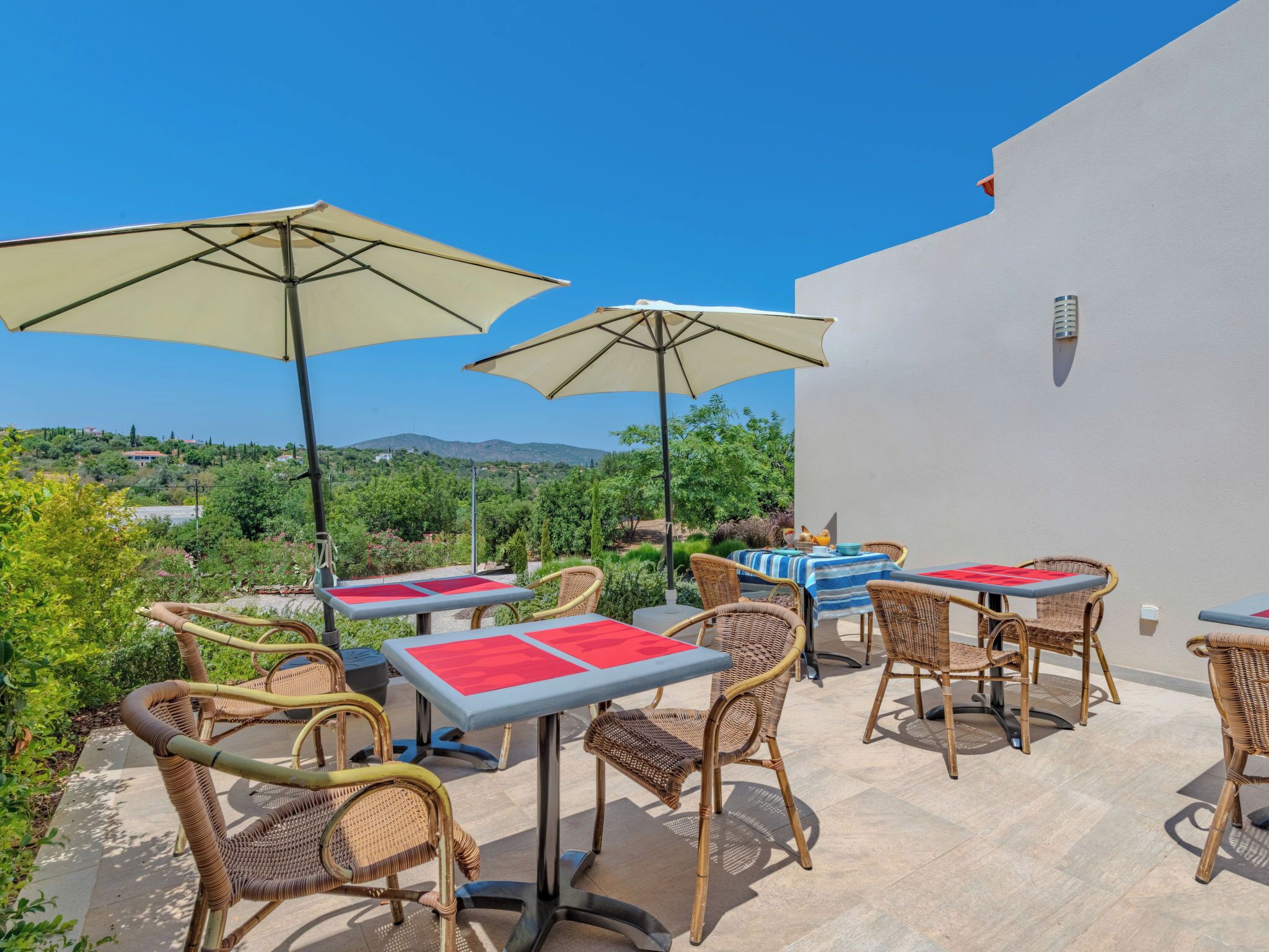 An outdoor patio with wicker chairs and tables, beige umbrellas, and a view of a hilly landscape.