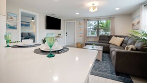 A modern living room with a white dining table, green glassware, a brown leather sofa, and a flat-screen TV mounted on the wall. - Calm Sea Cottage: New Renovation, Modern 2BR (Bradenton)
