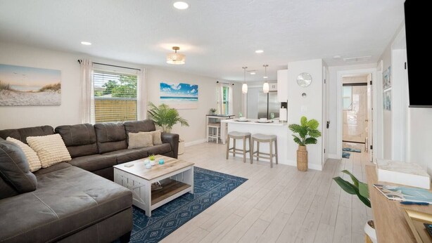 A modern living room with a brown leather sofa, a white coffee table, and a blue patterned rug. There is a kitchen area with a white countertop and bar stools. - Calm Sea Cottage: New Renovation, Modern 2BR (Bradenton)