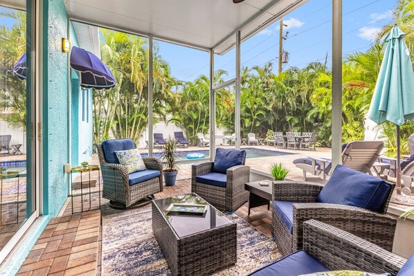 Un patio soleado con mobiliario de mimbre, una mesa de vidrio y vistas a una piscina y vegetación tropical.