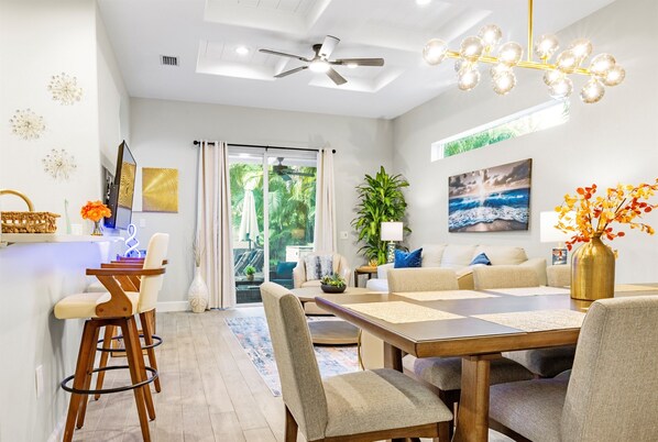 A modern living room with a dining area, featuring a ceiling fan, a chandelier, and a large window with curtains. - 2 Villas back to back, 2 Private Heated Pools, New Renovation (Sarasota)