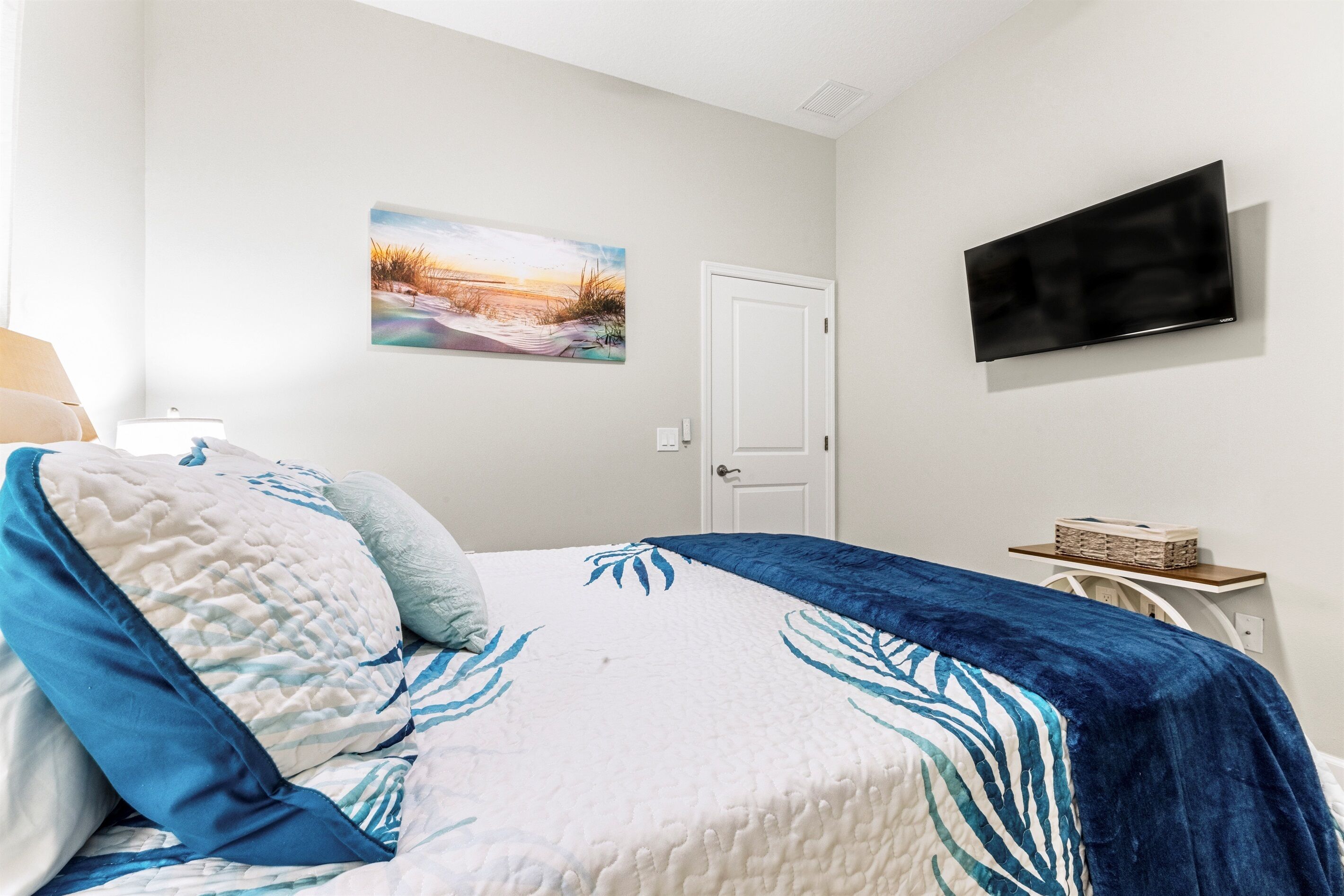 Une chambre à coucher avec un lit, un téléviseur fixé au mur, une porte et une petite table sur laquelle se trouve un panier.