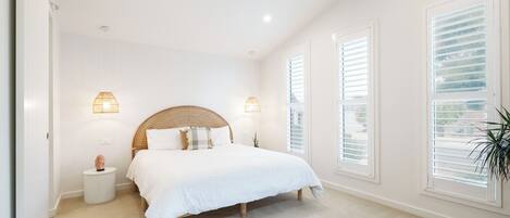 A bedroom with a large bed, two bedside lamps, a ceiling fan, and large windows with blinds.