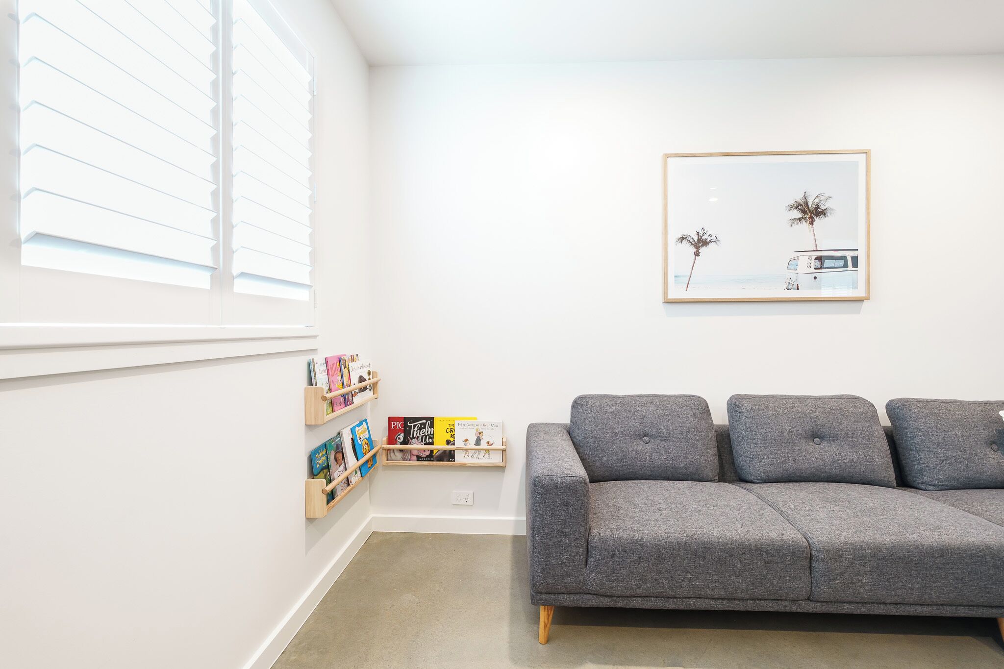Ein graues Sofa, ein Bücherregal mit Büchern, ein gerahmtes Bild von Palmen und ein Fenster mit Jalousien.