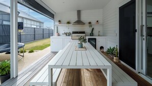 A modern outdoor kitchen with a white table, built-in appliances, and potted plants. - Large Retreat with Outdoor Living & Kids’ Paradise (Ocean Grove)