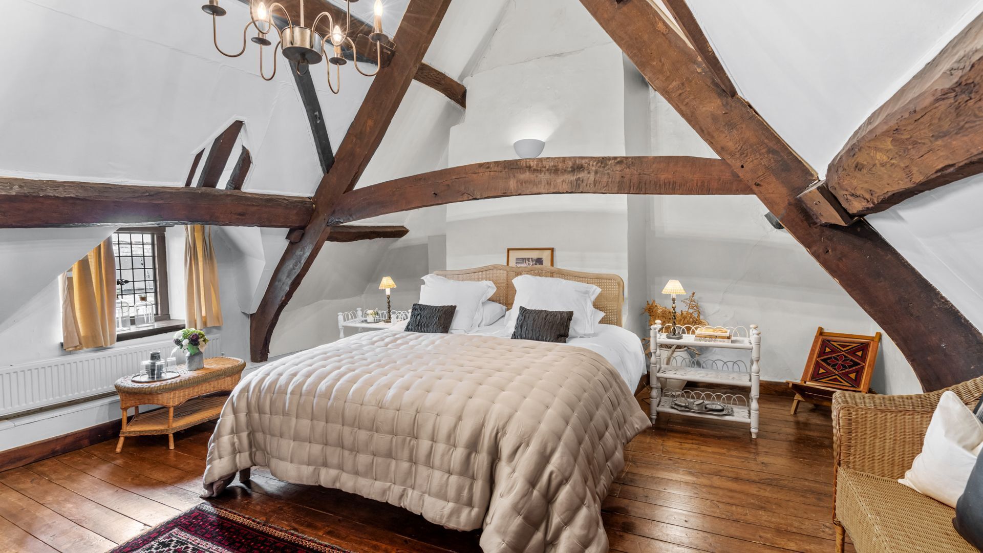 Une chambre avec un grand lit, des poutres en bois, un lustre, une fenêtre et un fauteuil en osier.