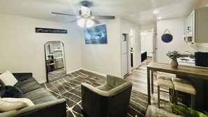 A modern living room with a sofa, a chair, a dining area with a table and chairs, a kitchen with a stove, and a ceiling fan. - Comfort Suite in charming Bay Shore (Bay Shore)