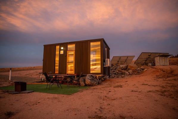 A small wooden cabin with a dining area, large windows, and solar panels.
