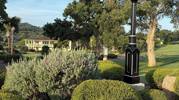 A clock with "Chloe Bay Resort" on it, surrounded by well-manicured bushes and trees, with a golf course and a building in the background.