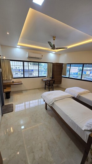A hotel room with two beds, a desk, a chair, a wardrobe, and a window with a view of buildings. - Hotel Priyanka palace lodging  (Jamner)
