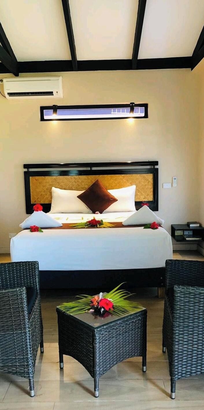 A hotel room with a bed, two chairs, a small table with flowers, an air conditioning unit, and a nightstand.
