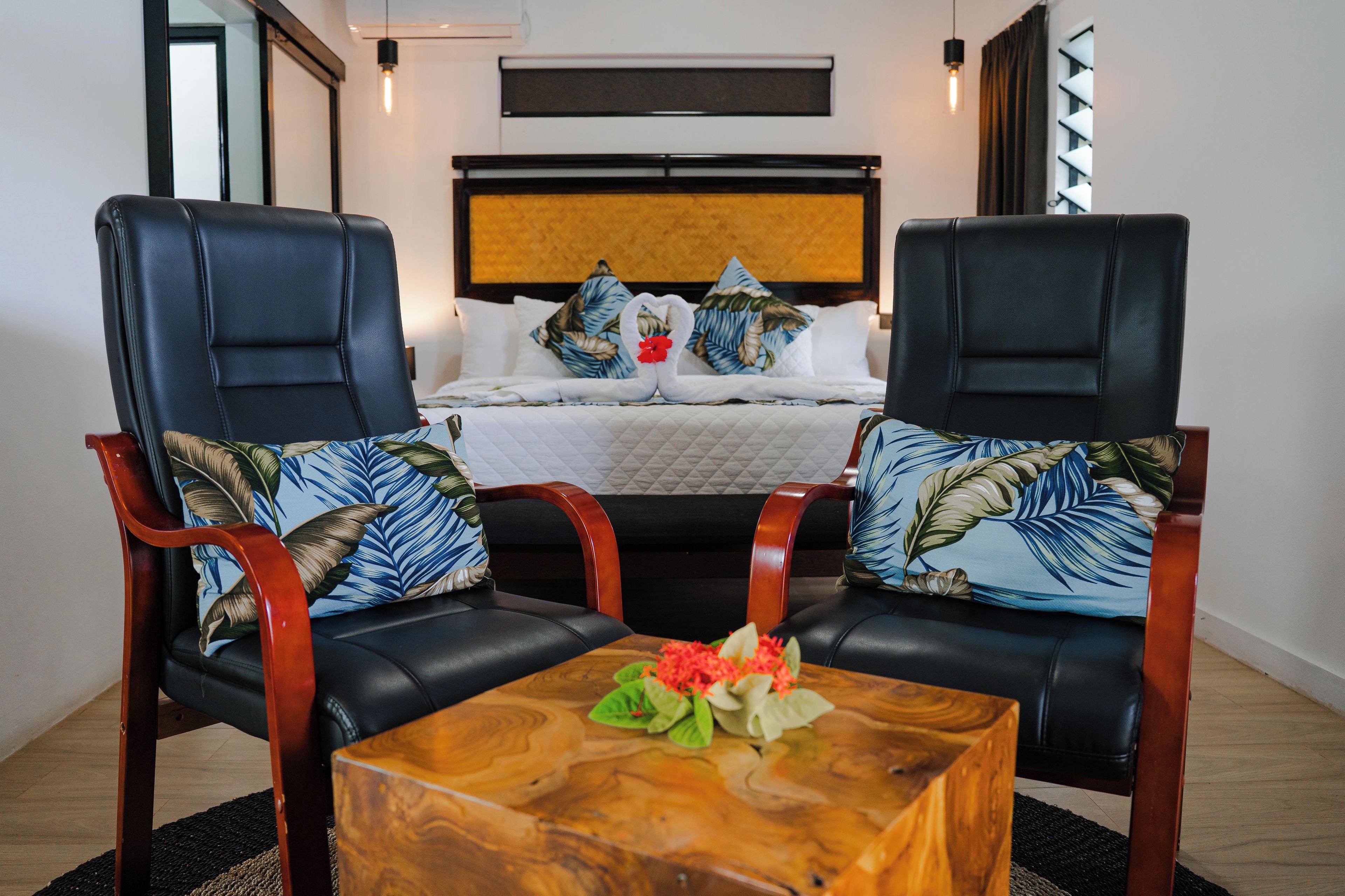A hotel room with a bed, two black leather chairs with tropical patterned cushions, a wooden coffee table with a flower arrangement, and a large painting on the wall.