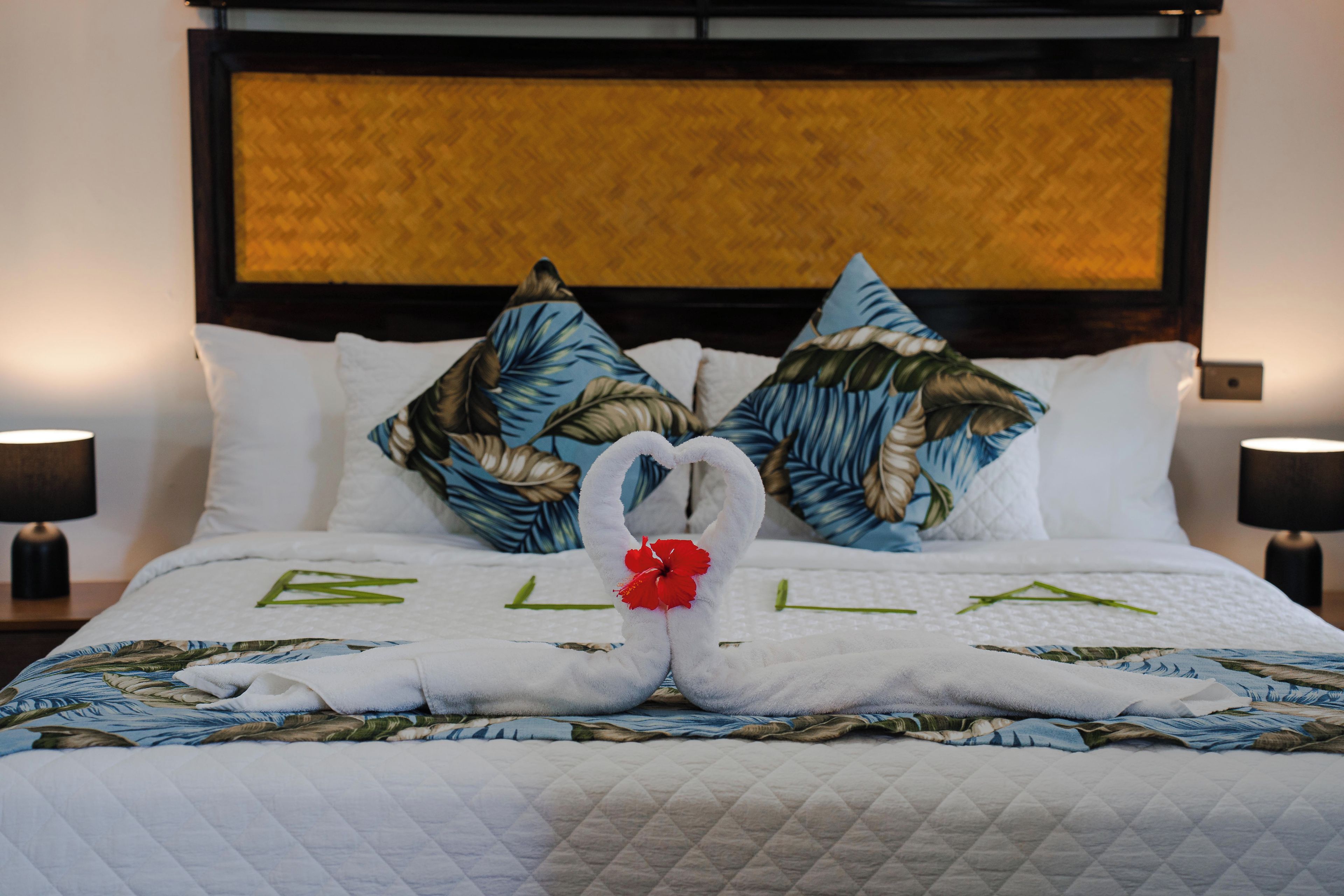 Una cama con colcha blanca, dos almohadas con estampado de hojas tropicales y dos toallas dispuestas en forma de cisne, adornadas con una flor roja.