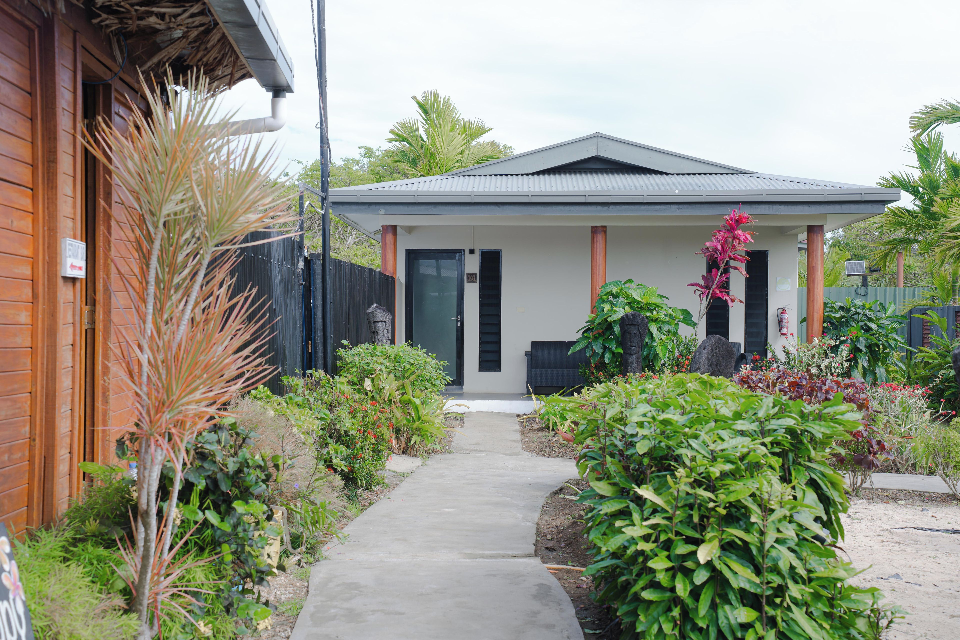 A residential property with a paved driveway leading to a front door, surrounded by various plants and a fenced garden area.