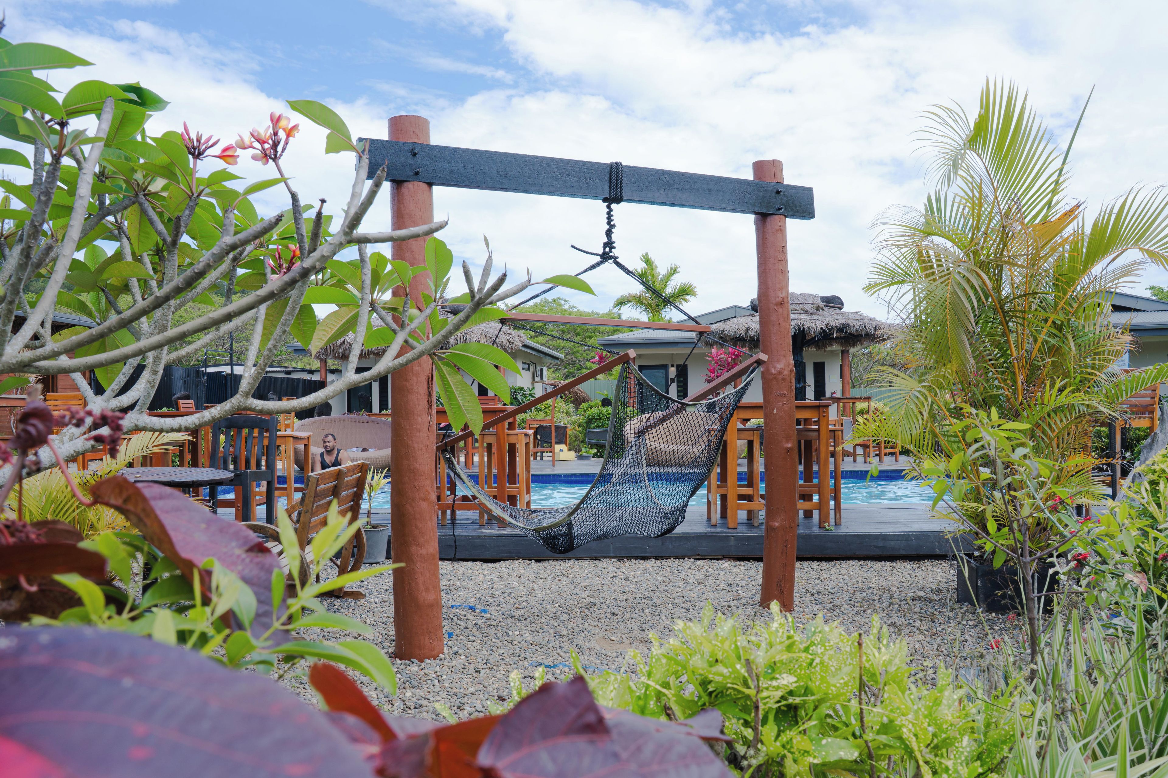 A hammock suspended over a pool area with a wooden deck, surrounded by tropical plants and a building with a thatched roof in the background.
