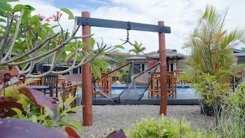 A hammock suspended over a pool area with a wooden deck, surrounded by tropical plants and a building with a thatched roof in the background.