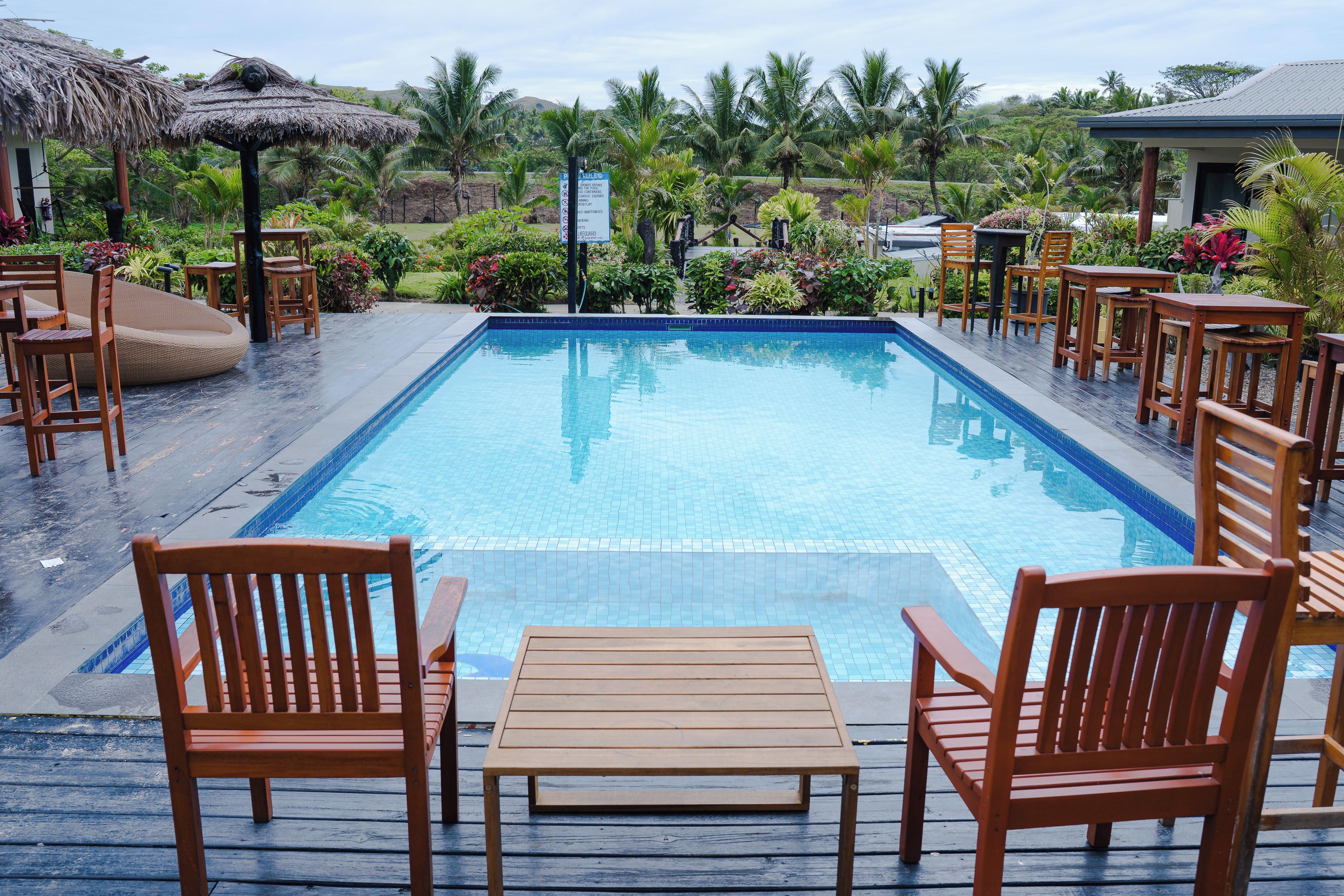 A swimming pool with wooden chairs and tables, surrounded by lush greenery and a thatched-roof structure.