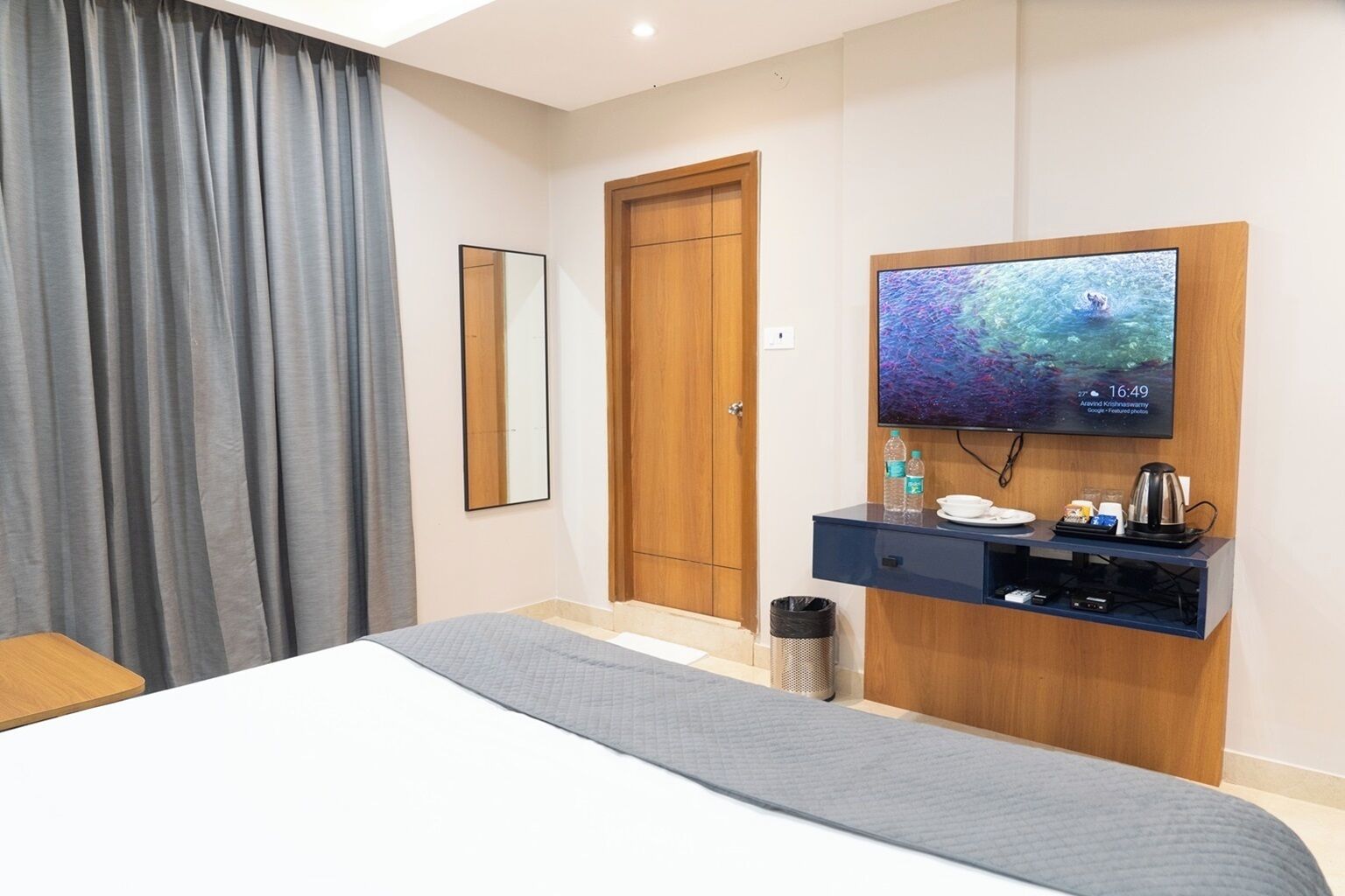 Habitación de hotel con cama, televisor montado en una pared de madera, una mesita con hervidor de agua y un cubo de basura.