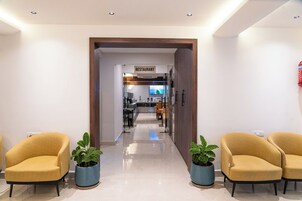 A modern hotel lobby with a reception desk, a restaurant sign, and two yellow armchairs with potted plants.