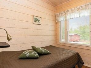 A room with wooden walls, a bed with a brown quilt, two green pillows, a framed picture on the wall, and a window with blinds.