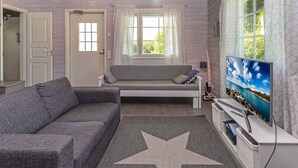 Uma sala de estar com um sofá cinzento, um tapete com padrão de estrelas, uma TV sobre um móvel e uma janela com cortinas estampadas.