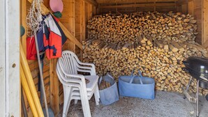 Interior de uma cabana de madeira com uma pilha de lenha, uma cadeira e uma churrasqueira.