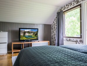 Une chambre avec un téléviseur posé sur un meuble en bois, deux lits avec une literie sombre, une commode blanche et une fenêtre donnant sur un paysage verdoyant.