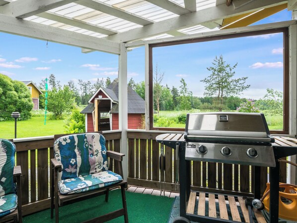 A covered outdoor area with a grill, a bench with a floral cushion, and a view of a house and greenery.