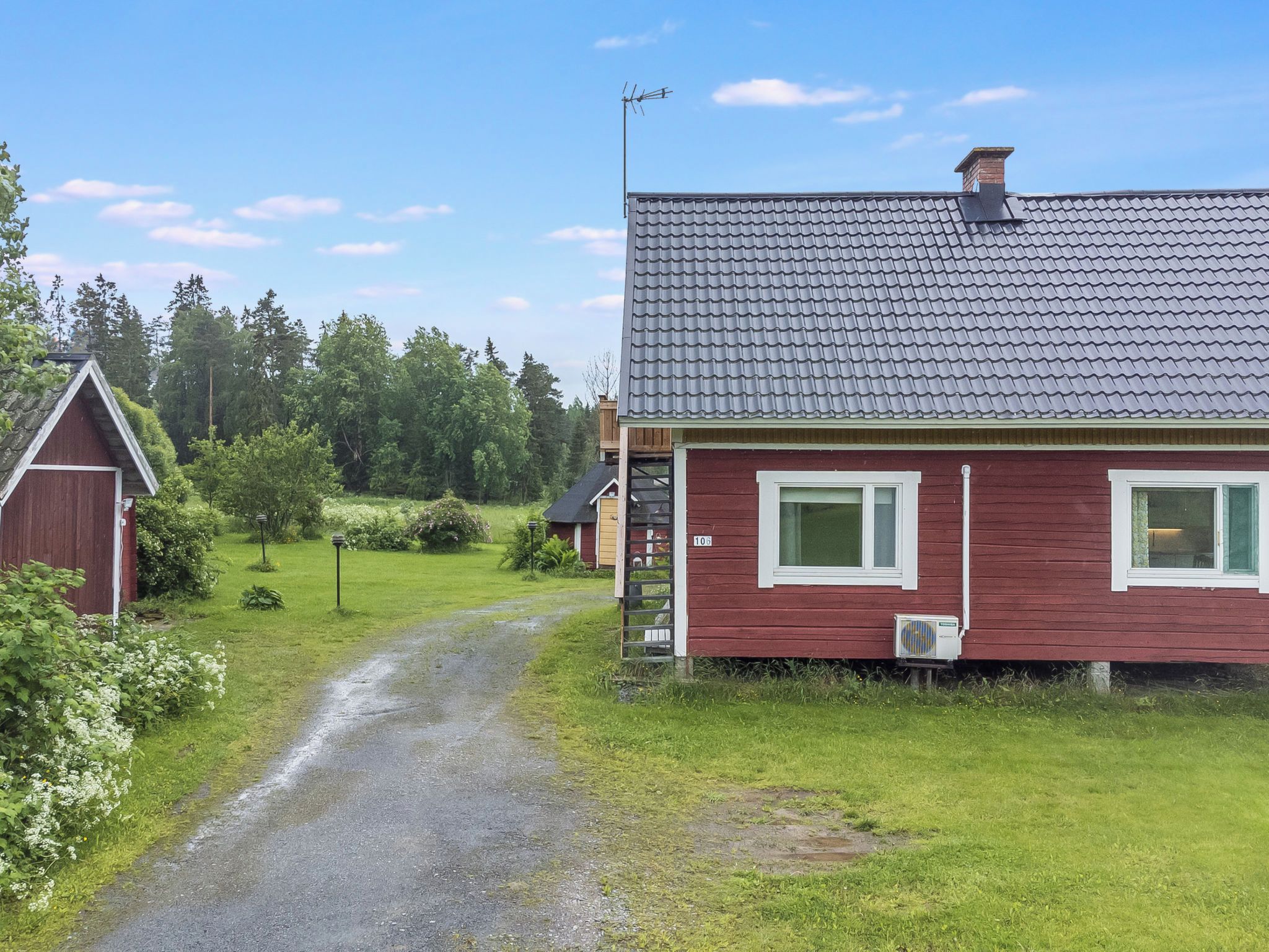 Punainen talo, jossa on harmaa katto, sorapäällysteinen ajotie ja puinen lato maaseutumaisemassa.