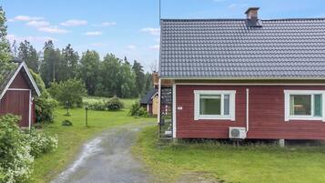 Punainen talo, jossa on harmaa katto, sorapäällysteinen ajotie ja puinen lato maaseutumaisemassa.