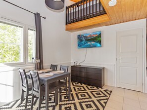 A dining area with a table, chairs, a patterned rug, a painting on the wall, and a window with curtains.