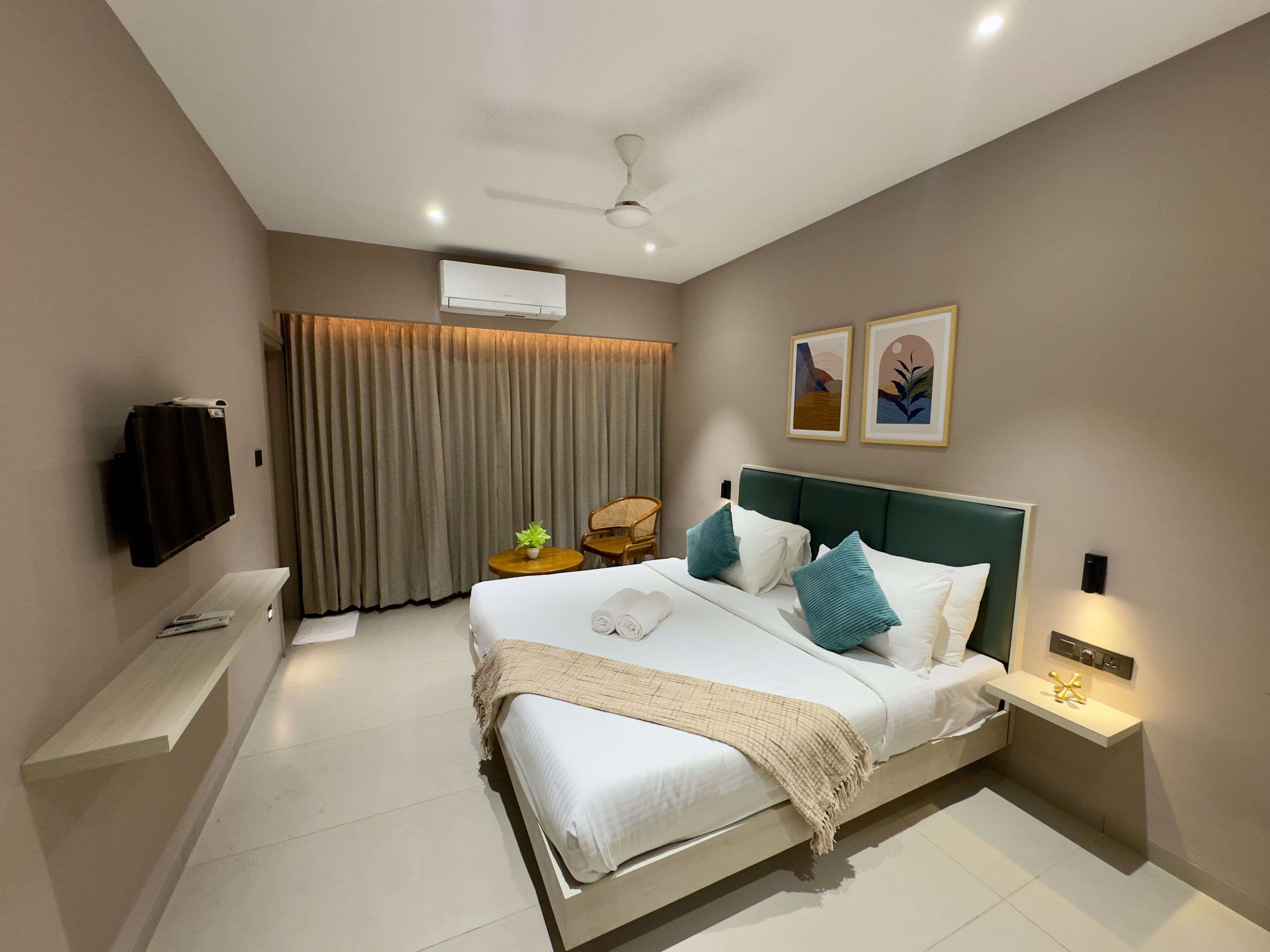 A modern hotel room with a large bed, a ceiling fan, a flat-screen TV, a small table with a plant, and a wall-mounted air conditioner.