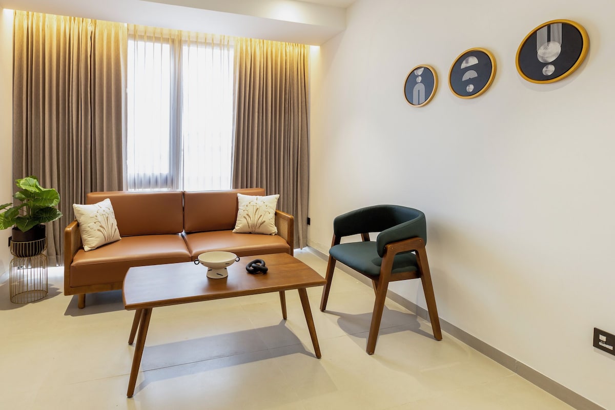 A modern living room with a brown sofa, a wooden coffee table, a green armchair, and a large window with curtains.