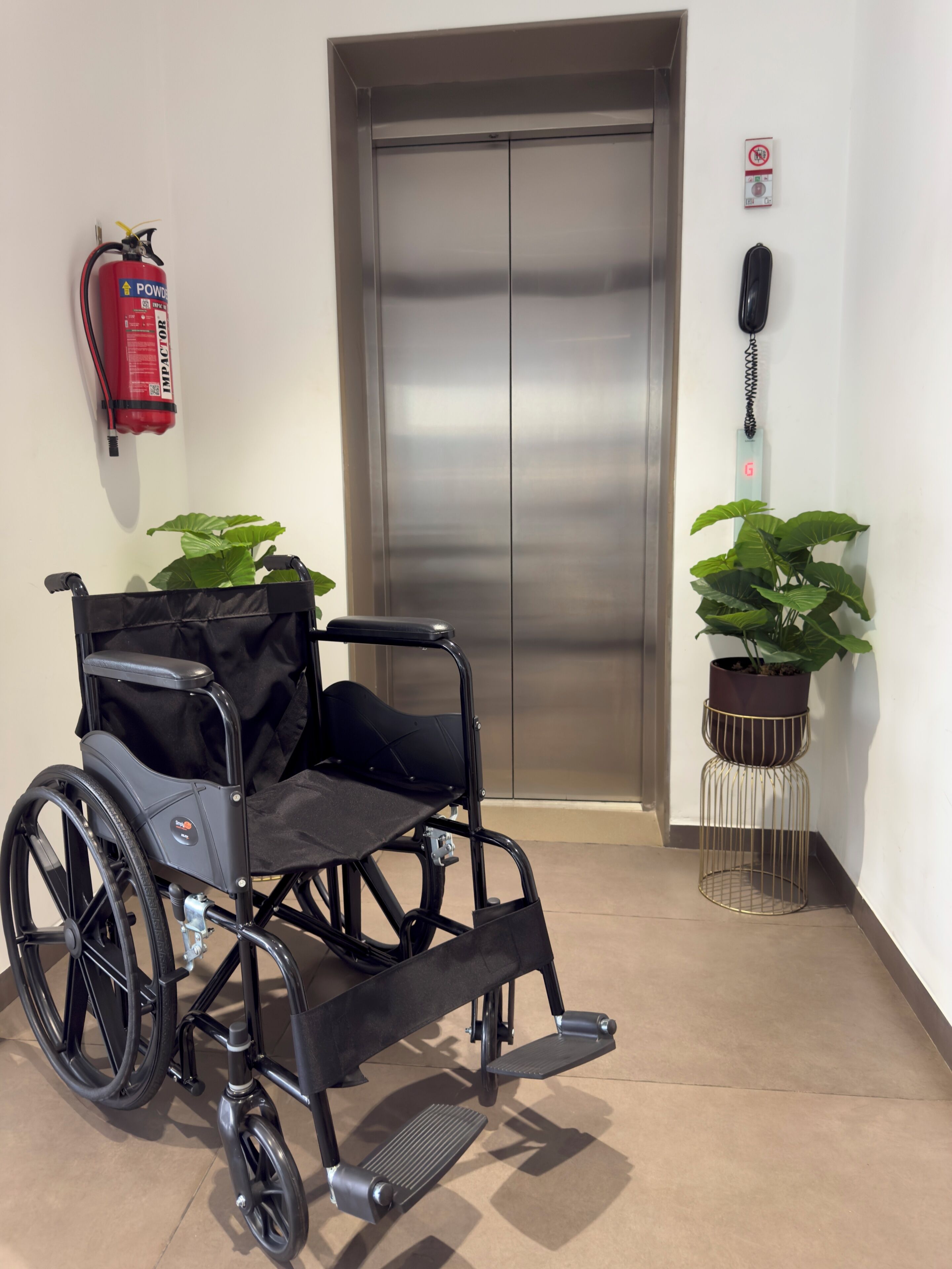 A wheelchair, a fire extinguisher, an elevator, and potted plants in a hallway.