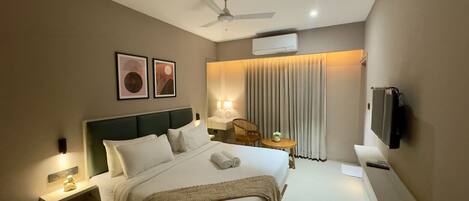 A modern hotel room with a large bed, a ceiling fan, a flat-screen TV, and a small table with a lamp.