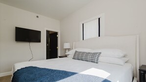 A bedroom with a large bed, a flat-screen TV mounted on the wall, a window, and a door.
