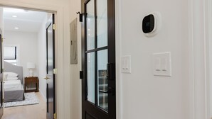 A hotel room with a bed, bedside table, lamp, and a digital thermostat on the wall.