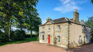 A stone house with a red door, surrounded by a gravel driveway and lush greenery.