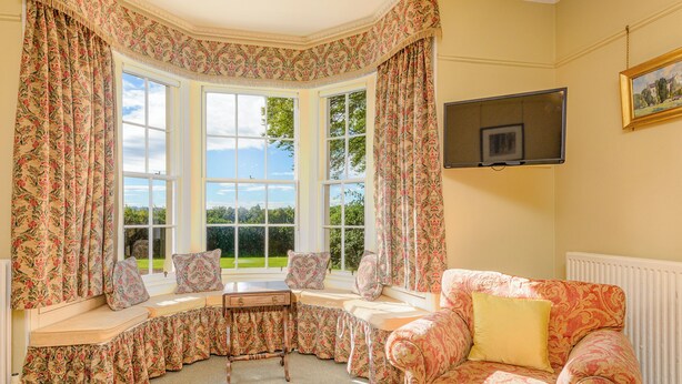 A room with a bay window, a sofa, a small table, and a television mounted on the wall.
