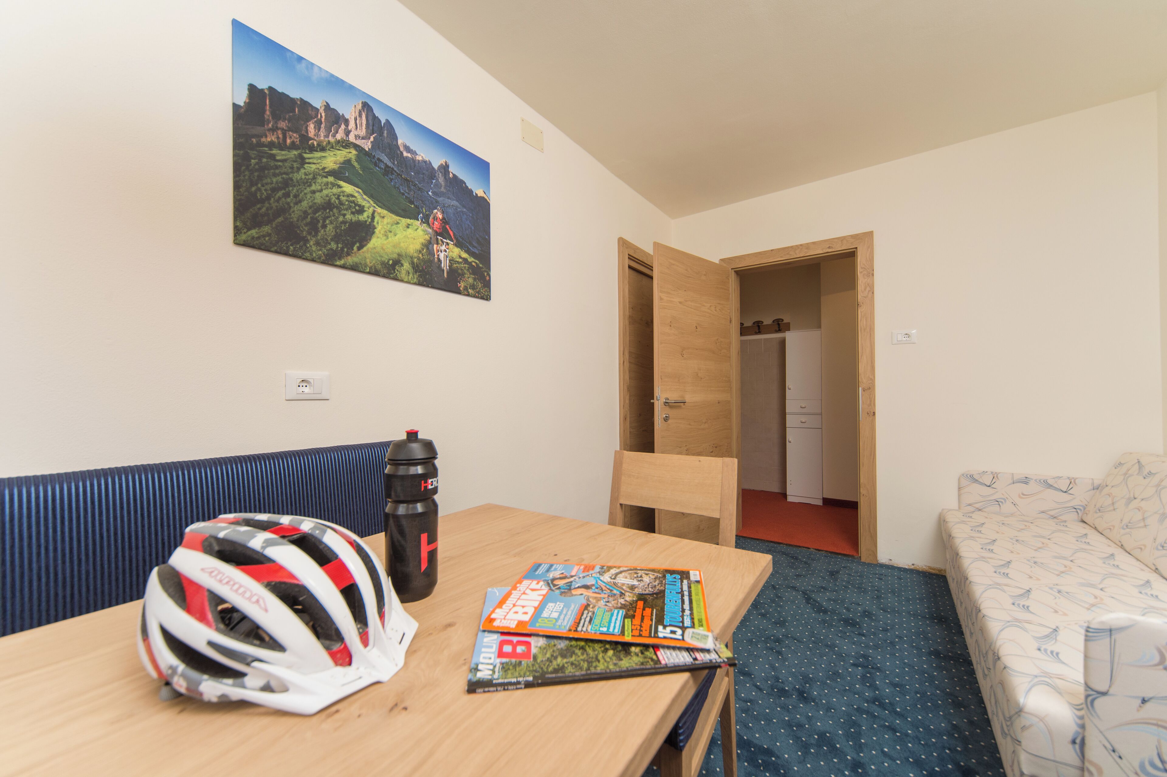 Quarto de hotel com mesa de madeira, uma revista, um capacete, garrafa de água, sofá e um quadro na parede.