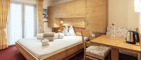 A hotel room with a wooden bed, a small table, a chair, and a kitchenette area.