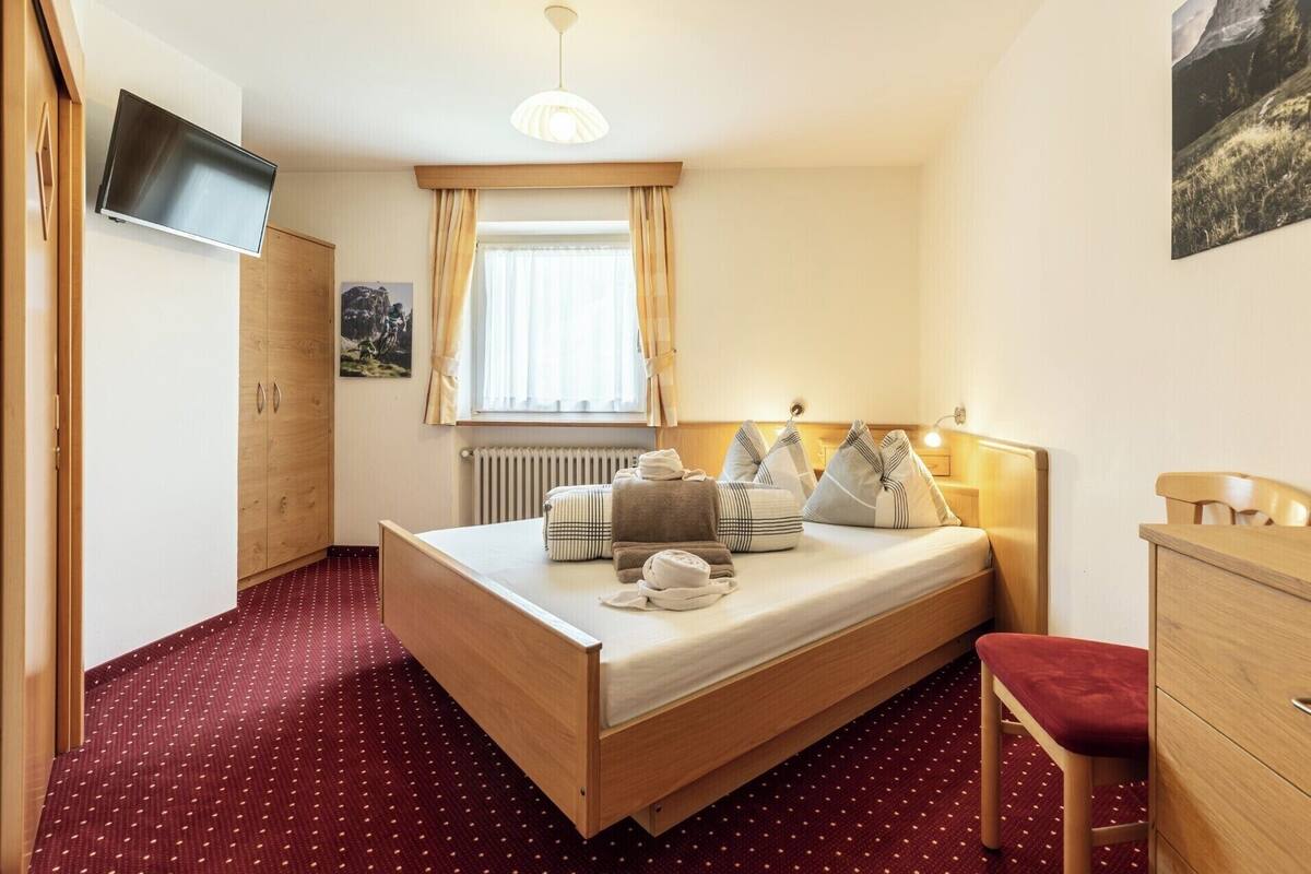 Un dormitorio con una cama, un aparador de madera, un televisor montado en la pared y un ventanal con cortinas.