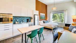 A modern kitchen with white cabinets, a wooden dining table, and green chairs. There is a bed with a view of trees through the window.