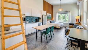 A modern kitchen with a wooden ladder, a dining area with green chairs, and a cozy living space with a bed and a window.