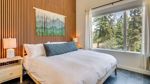 A bedroom with a large bed, wooden panel wall, a painting, a nightstand with a lamp, and a window with a view of trees.
