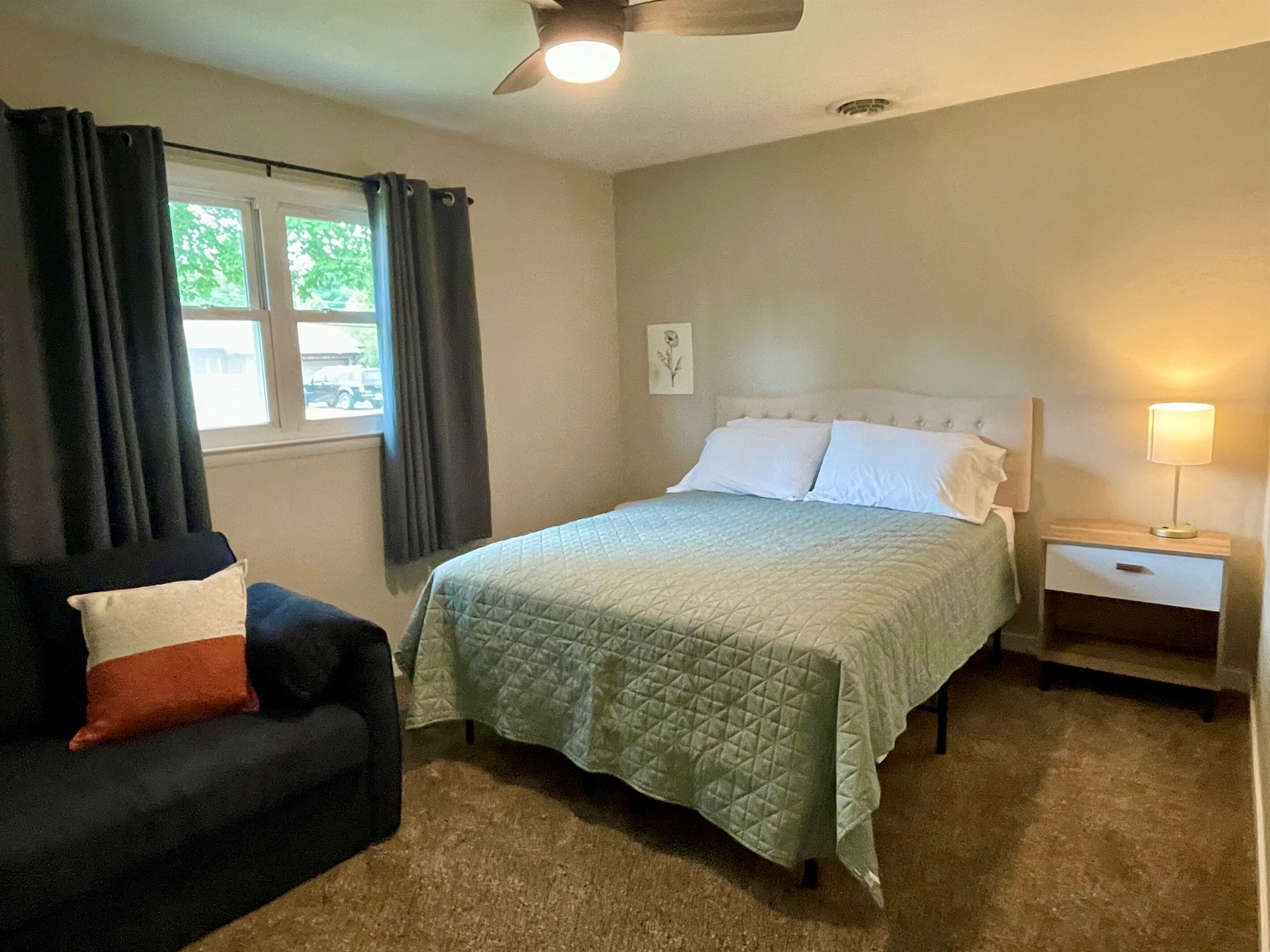 A bedroom with a bed, a nightstand, a lamp, a chair, and a window with curtains.